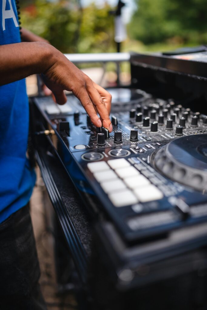 Close-up of a DJ's hand adjusting a mixer outdoors, with vibrant surroundings and equipment focus.